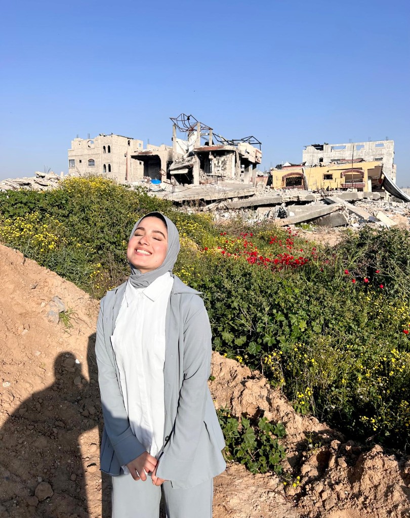Nour Elassy standing among flowers with destroyed buildings in Gaza behind her
